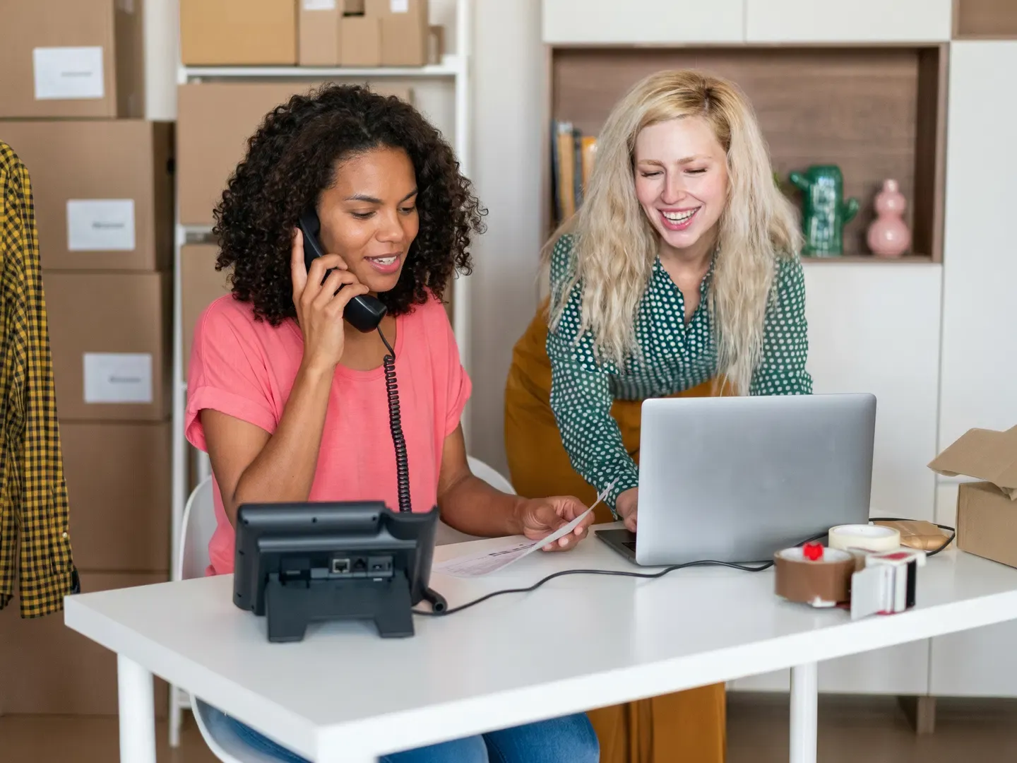 Two women managing orders at a desk using a desk phone and a laptop.